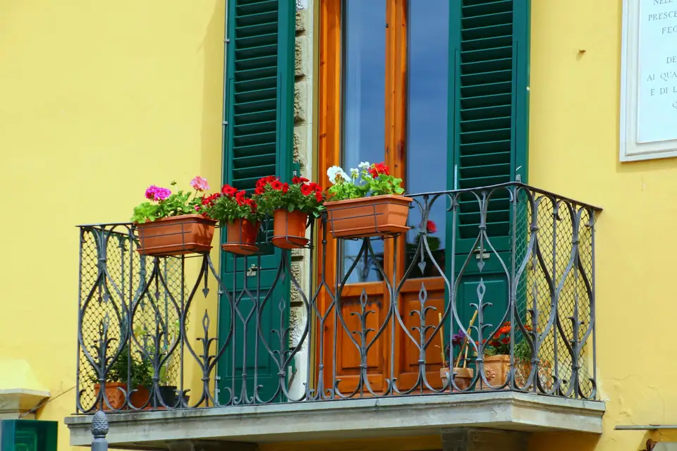 Terracotta window boxes of red and pink flowers on a wrought iron balcony with green shutters